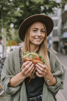 Smiling young woman with sandwich standing on street - MFF09029