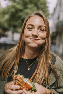 Lächelnde junge Frau mit aufgeblasenen Wangen, die ein Sandwich isst - MFF09030