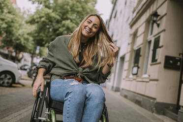 Happy woman sitting in wheelchair at sidewalk - MFF09031