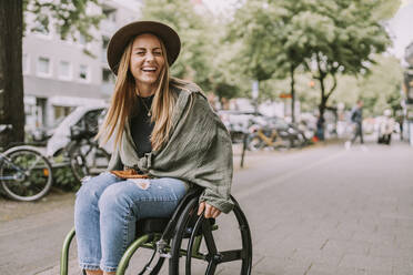 Cheerful young woman sitting in wheelchair at sidewalk - MFF09033