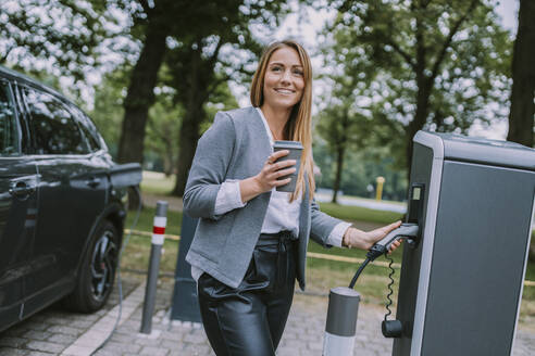 Frau mit Einwegbecher an der Ladestation für Elektrofahrzeuge - MFF09034