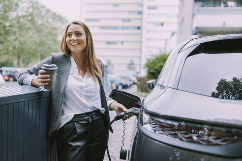 Glückliche Frau mit Einweg-Kaffeebecher, die ihr Auto an einer Tankstelle auflädt - MFF09035