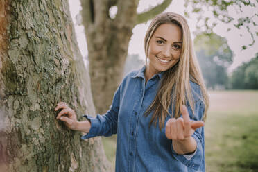 Lächelnde Frau gestikuliert bei einem Baum in einem öffentlichen Park - MFF09054