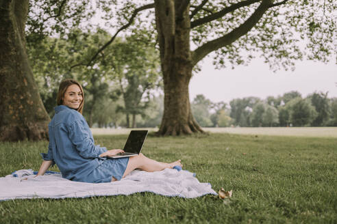 Lächelnde Frau mit Laptop auf einer Picknickdecke im Park sitzend - MFF09061