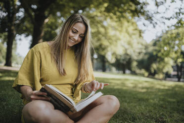 Lächelnde Frau, die im Gras sitzend in einem öffentlichen Park ein Buch liest - MFF09092