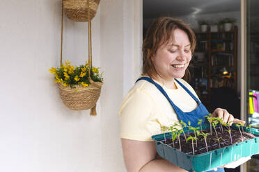 Happy woman with tray of tomato seedlings standing on balcony - IHF00779