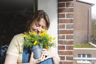 Lächelnde Frau mit geschlossenen Augen umarmt süßen Ginster Topfpflanze auf dem Balkon an einem sonnigen Tag - IHF00781