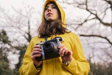 Young woman wearing yellow raincoat standing with camera in forest - AMWF00239