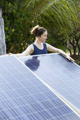 Woman standing behind solar panels under palm trees - NDF01419