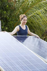 Smiling woman standing behind solar panels under palm trees - NDF01420