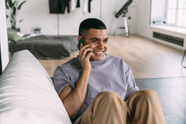 Smiling man talking on mobile phone sitting on sofa at home - VPIF05714