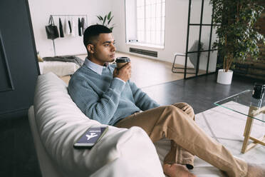 Mann hält Einweg-Kaffeebecher und sitzt neben einem Smartphone mit Flugmodus-Symbol auf dem Sofa zu Hause - VPIF05735