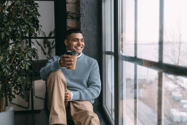 Glücklicher junger Mann mit Einweg-Kaffeebecher, der durch das Fenster schaut und zu Hause sitzt - VPIF05750