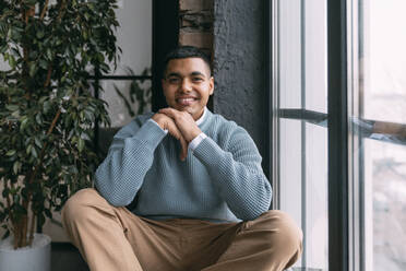 Smiling young man sitting by window at home - VPIF05752