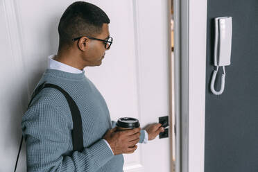 Junger Mann mit Brille, der eine Einweg-Kaffeetasse hält und die Tür öffnet - VPIF05757