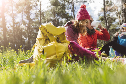 Glückliche Frau mit Rucksack sitzt bei Freunden essen und trinken im Wald an einem sonnigen Tag - JCCMF06085