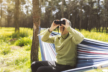 Man looking through binoculars sitting in hammock on sunny day - JCCMF06088