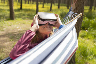 Lächelnde Frau mit Virtual-Reality-Simulator in der Hängematte liegend an einem sonnigen Tag - JCCMF06089