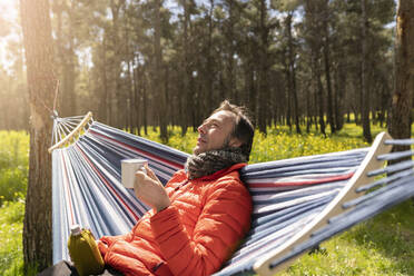 Mann hält Tasse in der Hängematte sitzend und genießt den sonnigen Tag - JCCMF06092