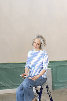 Frau auf Leiter sitzend mit Tablet-PC vor der Wand zu Hause - SEAF00750