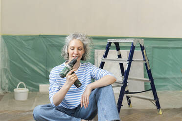 Smiling woman with drill gun sitting by ladder at home - SEAF00756