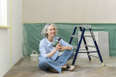 Happy woman with drill gun sitting by ladder at home - SEAF00758