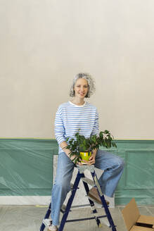 Glückliche Frau mit Topfpflanze, die zu Hause auf einer Leiter sitzt - SEAF00759
