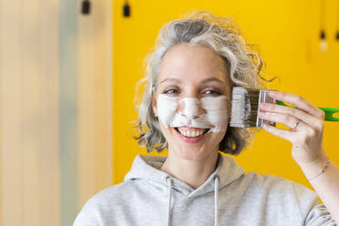 Smiling playful woman applying white paint on face with paintbrush - SEAF00776