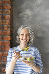 Glückliche Frau isst Salat beim Mittagessen vor einer Wand - SEAF00784