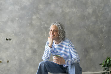 Glückliche Frau mit Hand am Kinn, die eine Tasse hält, sitzt vor einer Wand - SEAF00792