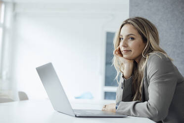Woman with hand on chin and laptop at desk in office - JOSEF08483