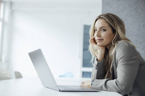 Frau mit Hand am Kinn und Laptop am Schreibtisch im Büro - JOSEF08483