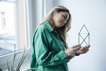 Smiling woman looking at house model standing in front of window - JOSEF08503