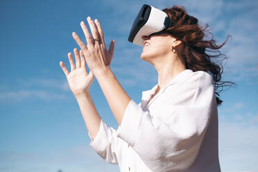 Frau mit Virtual-Reality-Simulator steht vor dem Himmel an einem sonnigen Tag - AMWF00249