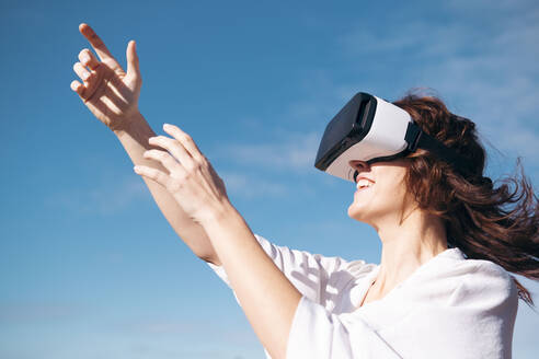 Glückliche Frau mit Virtual-Reality-Simulator vor dem Himmel an einem sonnigen Tag stehend - AMWF00250