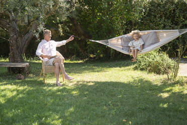 Senior man sitting by granddaughter enjoying hammock swing at garden - SVKF00114