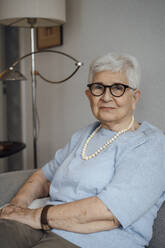 Smiling senior woman wearing eyeglasses sitting on sofa at home - JOSEF08553