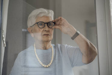 Senior woman wearing eyeglasses looking through window - JOSEF08574