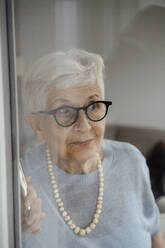 Curious senior woman wearing eyeglasses looking through glass window at home - JOSEF08576