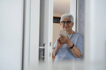 Glückliche ältere Frau mit Mobiltelefon, die zu Hause vor der Tür steht - JOSEF08577