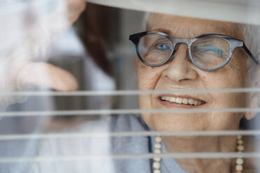 Happy senior woman looking through window blinds - JOSEF08581