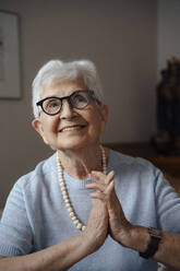 Happy senior woman with hands clasped sitting at home - JOSEF08587