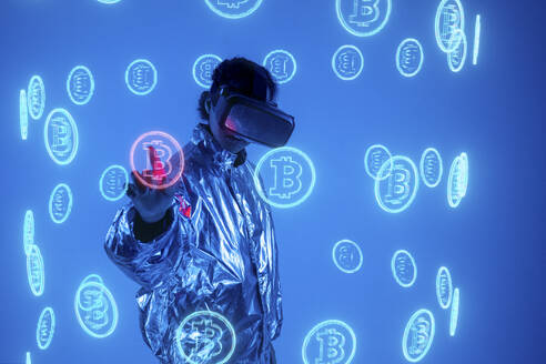Frau in Virtual-Reality-Simulator berührt Bitcoin-Symbol auf dem Bildschirm vor blauem Hintergrund - VPIF05774