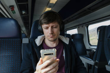 Mann mit Kopfhörern und Smartphone auf einem Sitz im Zug sitzend - TYF00129
