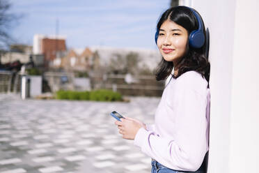 Lächelnde Frau mit Kopfhörern und Smartphone an der Wand lehnend - AMWF00265