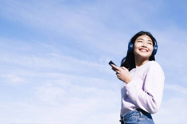 Lächelnde Frau mit Kopfhörern und Smartphone an einem sonnigen Tag - AMWF00272
