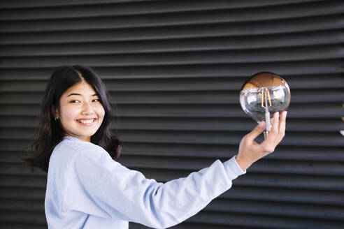 Glückliche junge Frau mit Glühbirne an einem Wellblechfensterladen - AMWF00279