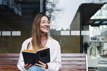 Glückliche Frau mit Buch auf einer Bank vor einer Glaswand sitzend - PNAF03660
