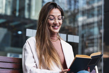 Lächelnde Frau mit Brille, die vor einer Glaswand sitzend ein Buch liest - PNAF03663