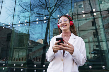 Lächelnde Frau mit drahtlosen Kopfhörern und Mobiltelefon vor einer Glaswand stehend - PNAF03682